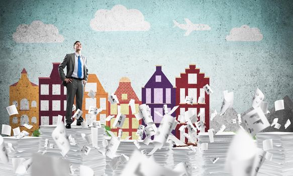 Confident businessman in suit standing on pile of documents among flying papers with drawn cityscape background. Mixed media.