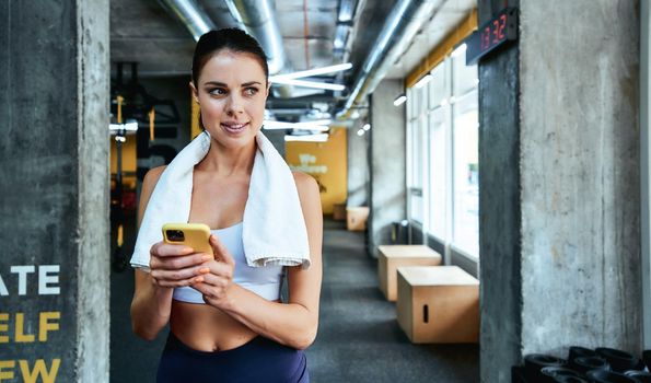 Texting sms. Young focused fitness woman in sportswear holding her smartphone and looking aside while exercising at gym. Sport, wellness and healthy lifestyle