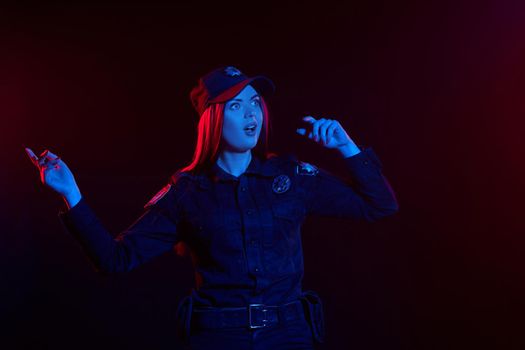 Close-up shot of a shocked redheaded girl police officer in a uniform and a cap looking away while posing against a black background with red and blue backlighting. Defender of citizens is ready to enforce a law and stop a crime.
