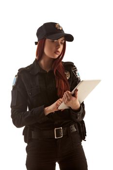 Lovely ginger policewoman in a black uniform and a cap is holding a tablet in her arms and typing something on it, isolated on white background. Defender of citizens is ready to enforce a law and stop a crime.