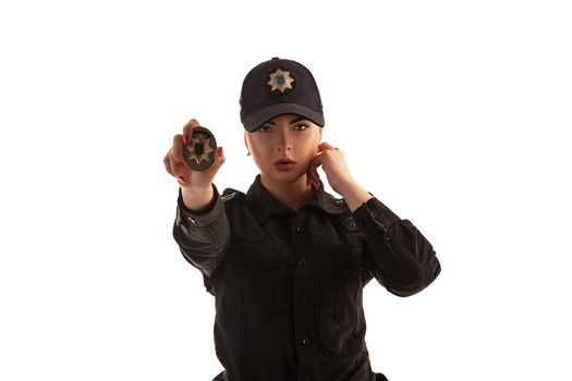 Close-up shot of a strong redheaded policewoman in a black uniform and a cap showing her badge and looking at the camera, isolated on white background. Defender of citizens is ready to enforce a law and stop a crime.