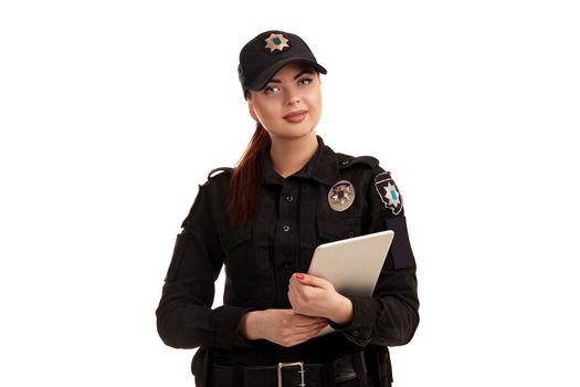 Close-up shot of a cute redhead policewoman in a black uniform and a cap holding a tablet in her arms. She is looking at the camera and smiling while posing isolated on white background. Defender of citizens is ready to enforce a law and stop a crime.