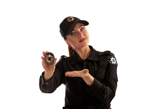 Close-up shot of a charming ginger policewoman in a black uniform and a cap showing her badge and looking at the camera, isolated on white background. Defender of citizens is ready to enforce a law and stop a crime.