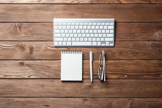 Still life of office workspace with supplies. Flat lay old wooden desk with white computer keyboard, pen and notebook. Top view business concept. Vintage brown wooden surface with copy space.