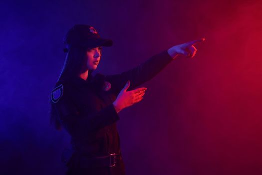 Charming redheaded female traffic controller in a uniform and a cap is regulating traffic with signals posing sideways against a black background with red and blue backlighting. Defender of citizens is ready to enforce a law and stop a crime.