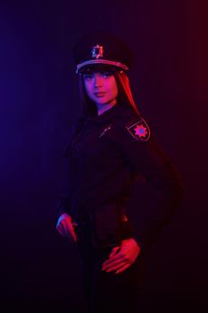 Young redheaded maiden police officer in a uniform and a cap, with bright make-up is looking at the camera and posing sideways against a black background with red and blue backlighting. Defender of citizens is ready to enforce a law and stop a crime.