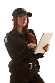 Nice redhead policewoman in a black uniform and a cap is holding a tablet in her arms, typing something on it and looking at the camera, isolated on white background. Defender of citizens is ready to enforce a law and stop a crime.