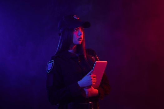 Beautiful redheaded woman police officer in a uniform and a cap, with bright make-up is holding her tablet and posing sideways against a black background with red and blue backlighting. Defender of citizens is ready to enforce a law and stop a crime.