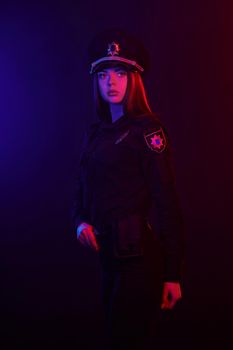 Young redheaded lady police officer in a uniform and a cap, with bright make-up is looking at the camera and posing sideways against a black background with red and blue backlighting. Defender of citizens is ready to enforce a law and stop a crime.