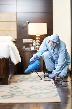 Man in a protective clothes spraying disinfectants near bed in hotel room. Coronavirus and quarantine concept