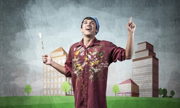 Young male artist holding paintbrush. Happy painter in shirt and bandana standing on background colorful picture on wall. Modern cityscape with high skyscrapers artwork. Creative hobby and profession