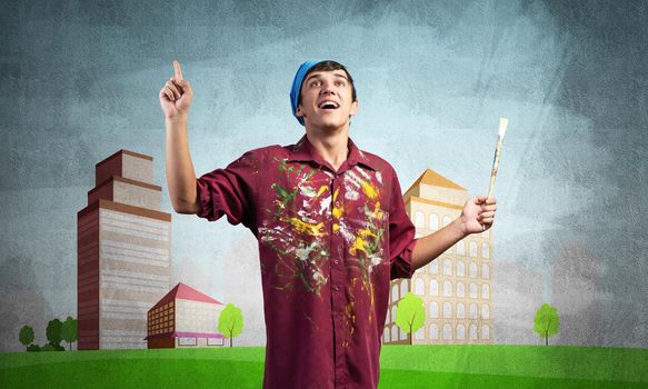 Young male artist holding paintbrush. Happy painter in shirt and bandana standing on background colorful picture on wall. Modern cityscape with high skyscrapers artwork. Creative hobby and profession