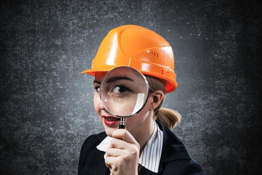 Building inspector looking through magnifying glass. Young construction specialist in safety helmet with magnifier on grey wall background. Consultancy and expertise. Professional valuation services