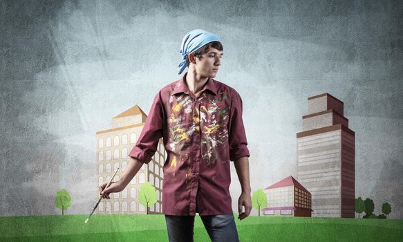 Young male artist holding paintbrush. Painter in shirt and bandana standing on background colorful picture on wall. Modern cityscape with high skyscrapers artwork. Creative hobby and profession