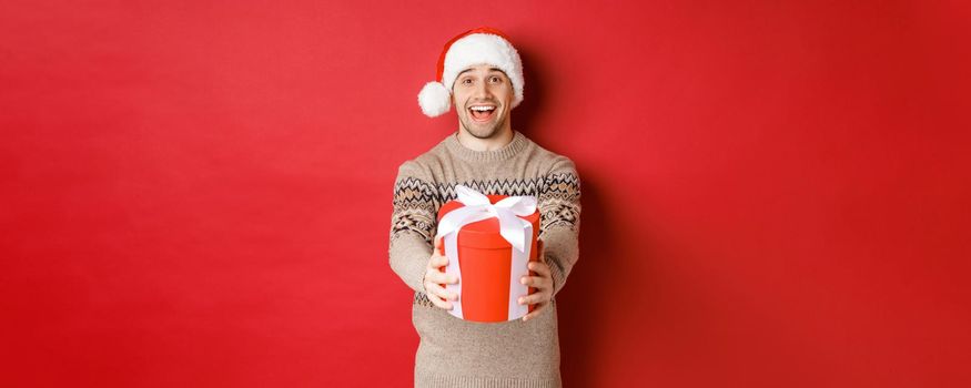 Image of happy man saying merry christmas and giving you a gift in box, smiling cheerful, celebrating winter holidays in santa hat and xmas sweater, standing over red background.