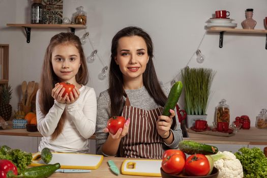 Happy loving family are cooking together. Delightful mother and her little princess are posing with tomato and cucumber at the kitchen, against a white wall with shelves and bulbs on it. Homemade food and little helper.