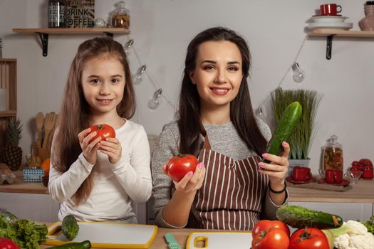 Happy loving family are cooking together. Delightful mommy and her little girl are posing with tomato and cucumber at the kitchen, against a white wall with shelves and bulbs on it. Homemade food and little helper.