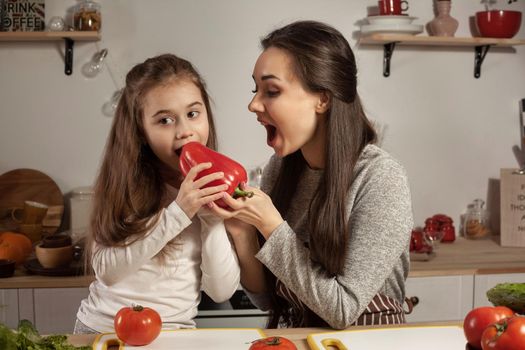 Happy loving family are cooking together. Loving mommy and her little girl are making a vegetable salad and tasting a red pepper at the kitchen, against a white wall with shelves and bulbs on it. Homemade food and little helper.