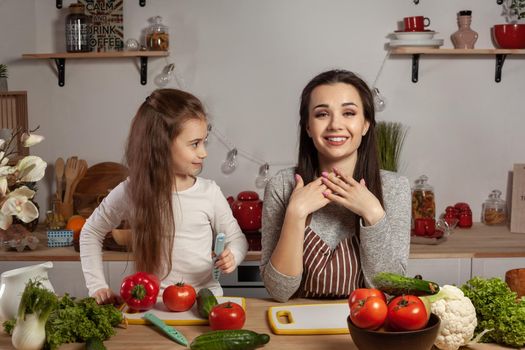 Happy loving family are cooking together. Lovely mother and her little princess are making a vegetable salad and having fun at the kitchen, against a white wall with shelves and bulbs on it. Homemade food and little helper.