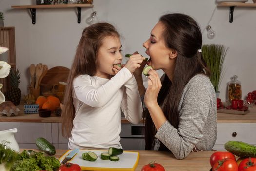 Happy loving family are cooking together. Alluring mother and her daughter are making a vegetable salad and tasting a cucumber at the kitchen, against a white wall with shelves and bulbs on it. Homemade food and little helper.