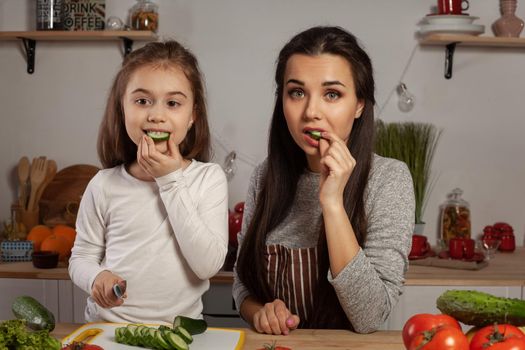 Happy loving family are cooking together. Alluring mother and her little princess are making a vegetable salad and tasting a cucumber at the kitchen, against a white wall with shelves and bulbs on it. Homemade food and little helper.