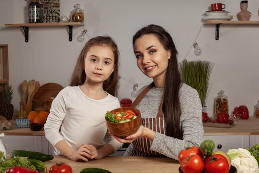 Happy loving family are cooking together. Smiling mum and her child are making a vegetable salad and looking at the camera at the kitchen, against a white wall with shelves and bulbs on it. Homemade food and little helper.