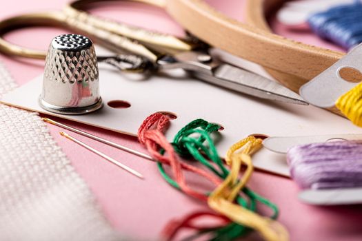 Embroidery set fot cross stitching. White fabric, thimble, embroidery hoop, colorful threads, scissors and needls. On pink background.