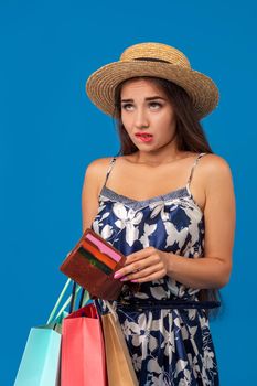 Portrait of unhappy young woman looking in her wallet in shopping center, spent too much, not enough cash, lost money, broke. Studio shot on blue background. Copy space
