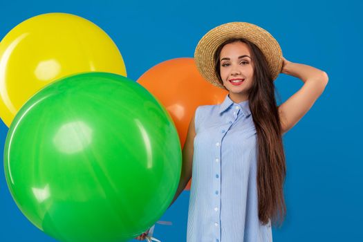 Cute brunette standing in a studio holding orange, yellow and green balloons. She wears light blue dress and has long and wavy hair. Celebration. Surprise