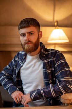 Portrait handsome bearded man wearing plaid shirt at modern cafe. Guy sitting in wooden chair and relaxing.