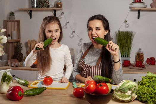 Happy loving family are cooking together. Wonderful mommy and her little girl are tasting cucumbers and having fun at the kitchen, against a white wall with shelves and bulbs on it. Homemade food and little helper.