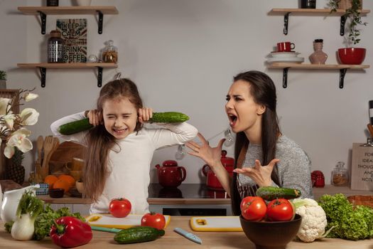 Happy loving family are cooking together. Brunette mom and her kid are making a vegetable salad and quarrelling at the kitchen, against a white wall with shelves and bulbs on it. Homemade food and little helper.