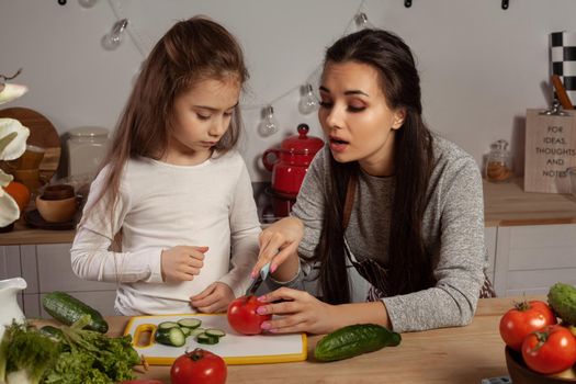 Happy loving family are cooking together. Nice mum and her child are making a vegetable salad and having fun at the kitchen, against a white wall with shelves and bulbs on it. Homemade food and little helper.