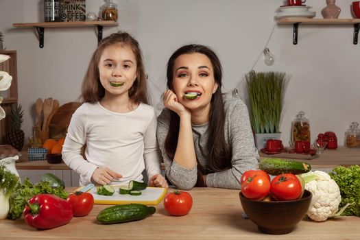 Happy loving family are cooking together. Attractive mum and her child are making a vegetable salad and tasting a cucumber at the kitchen, against a white wall with shelves and bulbs on it. Homemade food and little helper.