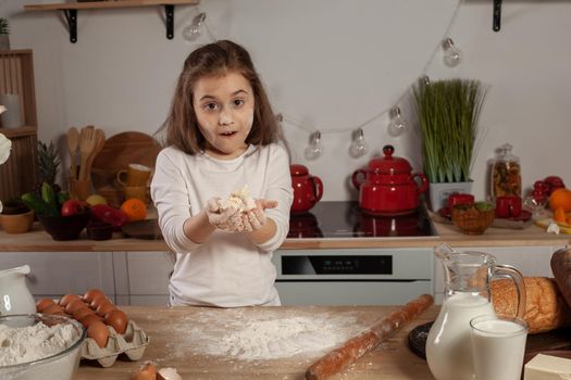 Charming little brunette lady dressed in a white blouse is showing a dough for baking a bread and looking at the camera at a kitchen against a white wall with shelves and bulbs on it. Happy childhood concept.