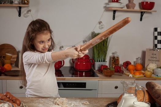 Beautiful little brunette lady dressed in a white blouse is playing with a rolling pin at a kitchen against a white wall with shelves and bulbs on it. Happy childhood concept.