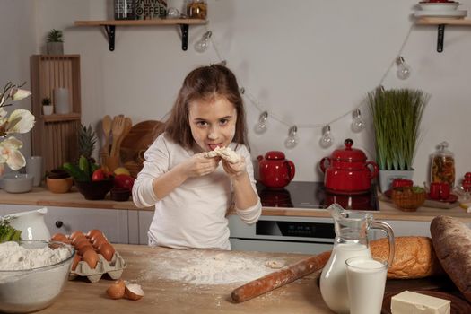 Charming little brunette child dressed in a white blouse is tasting the dough for baking a bread at a kitchen against a white wall with shelves and bulbs on it. Happy childhood concept.