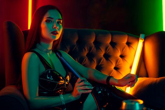 Lovely brunette model with a perfect make-up, in a sparkling necklace, leather dress and patent boots is smoking a hookah sitting sideways on an arm-chair at a luxury night club. Concept of pleasant spending time. Dark background with colorful neon backlights.