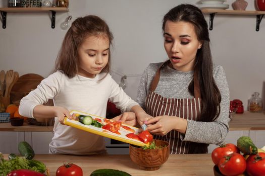 Happy loving family are cooking together. Beautiful mom and her kid are making a vegetable salad and having fun at the kitchen, against a white wall with shelves and bulbs on it. Homemade food and little helper.