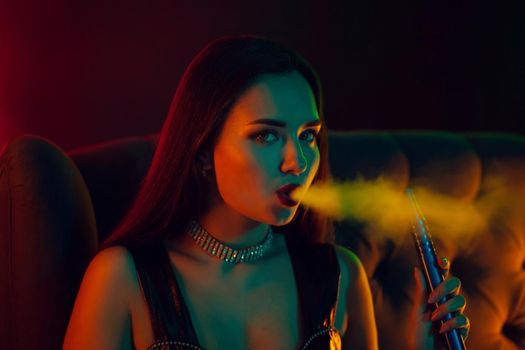 Close-up shot of a dazzling brunette female with a perfect make-up, in a sparkling necklace and a leather dress smoking a hookah exhaling a smoke sitting sideways on an arm-chair at a luxury night club. Concept of pleasant spending time. Dark background with colorful neon backlights.