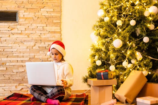 Little girl in a sweater for the New Year. A girl and a laptop at the Christmas tree with a book at Christmas. A child and a computer for the New Year.