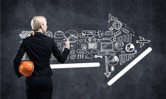 Businesswoman draws arrows consists of business doodles on blackboard. Back view woman with safety helmet and marker pen in hands. Idea generation and business development. Coaching and mentoring.