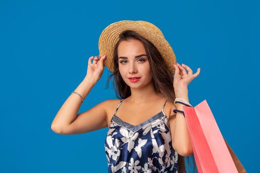 Portrait of a young woman in summer hat holding shopping bags and looking at the camera isolated over blue background. The concept of shopping