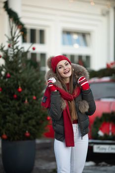 Pleasant fashionable winter girl smiling posing at red vintage car surrounded by snowflakes medium shot. Beautiful young woman enjoying december Christmas holiday having positive emotion