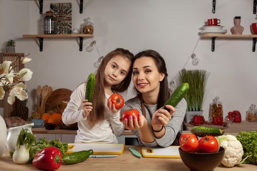 Happy loving family are cooking together. Good-looking mom and her kid are posing with tomato and cucumber and smiling at the kitchen, against a white wall with shelves and bulbs on it. Homemade food and little helper.