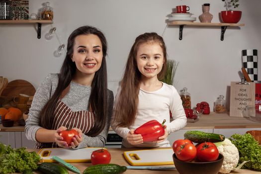 Happy loving family are cooking together. Loving mother and her daughter are making a vegetable salad and having fun at the kitchen, against a white wall with shelves and bulbs on it. Homemade food and little helper.