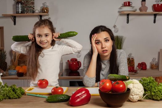 Happy loving family are cooking together. Brunette mum and her child are making a vegetable salad and tired of each other at the kitchen, against a white wall with shelves and bulbs on it. Homemade food and little helper.