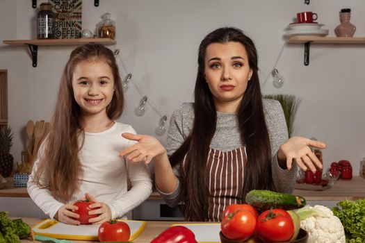 Happy loving family are cooking together. Lovely mother and her daughter are making a vegetable salad and tired of each other at the kitchen, against a white wall with shelves and bulbs on it. Homemade food and little helper.