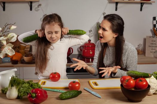 Happy loving family are cooking together. Brunette mother and her daughter are making a vegetable salad and quarrelling at the kitchen, against a white wall with shelves and bulbs on it. Homemade food and little helper.