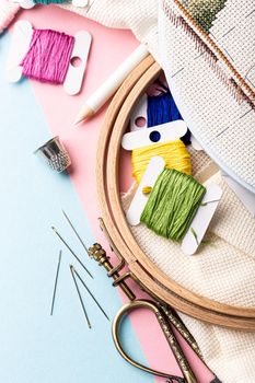 Overhead shot of embroidery set fot cross stitching. White fabric, embroidery hoop, colorful threads, scissors and needls. On pink blue background. Hobbies concept with copy space.